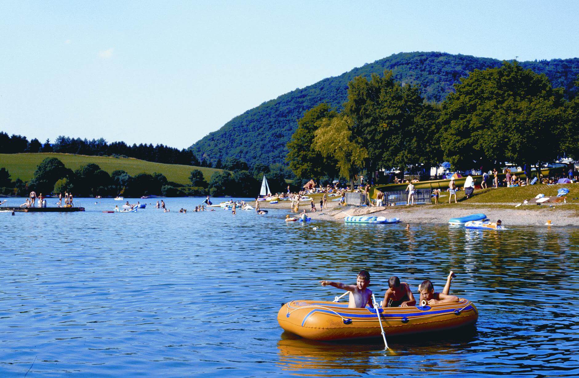 Der Diemelsee und das Strandbad im Sommer. Im Vordergrund drei Jungen in einem gelben Schlauchboot. Im Hintergrund der See in dem viele Menschen baden.