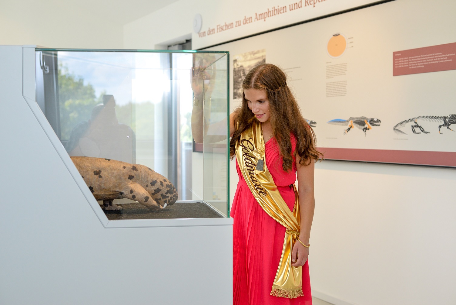 Die Goldmarie, eine Frau im roten Kleid und goldener Schärpe begutachtet ein Ausstellungsexemplar in einem Glaskasten.