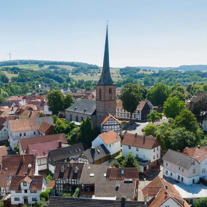 Die sommerliche Stadtansicht von Gemünden mit einigen Fachwerkhäusern, mittig die Kirche. Im Hintergrund eine grüne Landschaft.