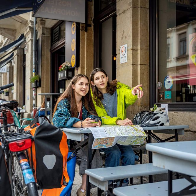 Zwei Radfahrerinnen sitzen draußen an einem Tisch, vor ihnen eine Radkarte, neben ihnen ein Fahrrad mit Satteltaschen