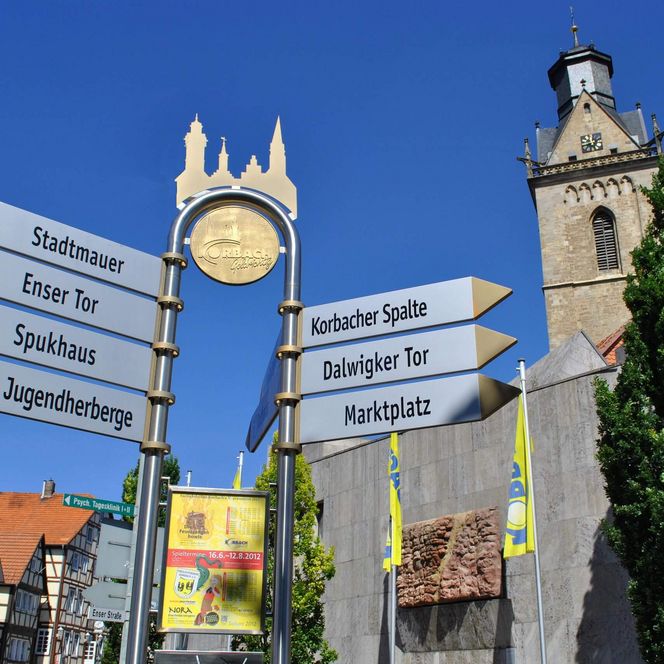 Im Bildmittelpunkt sieht man vor einem blauen Himmel zahlreiche Hinweisschilder, die zu den zahlreichen Sehenswürdigkeiten in der Hansestadt Korbach weisen. Im Hintergrund der Kirchturm der Kilianskirche, davor das Wolfgang-Bonhage-Museum.