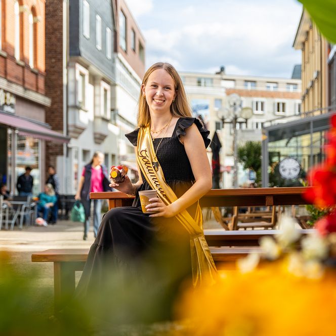 Die Korbacher Goldmarie sitzt in der Fußgängerzone, hält Kaffee und Muffin in den Händen und lächelt in die Kamera. Sie trägt ein schwarzes Kleid mit goldener Schärpe.