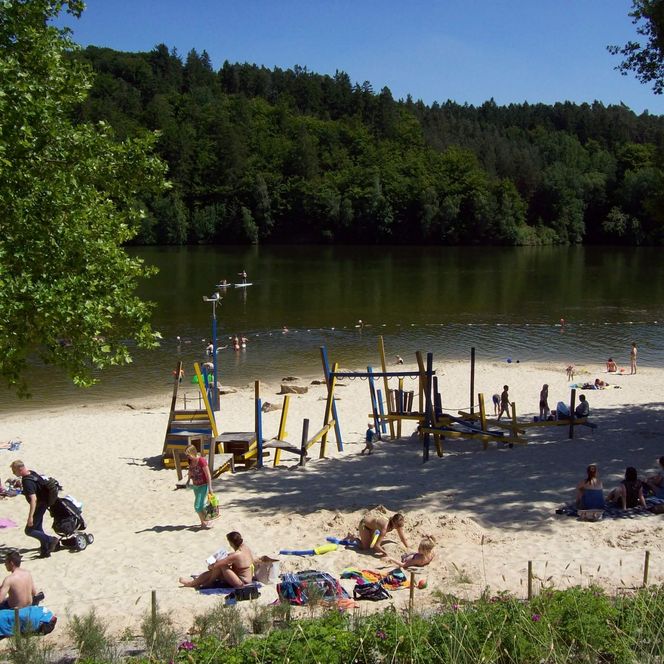 Der belebte Sandstrand mit Klettergerüst am Strandbad im Sommer. Im Hintergrund der Twistesee unter blauem Himmel. 