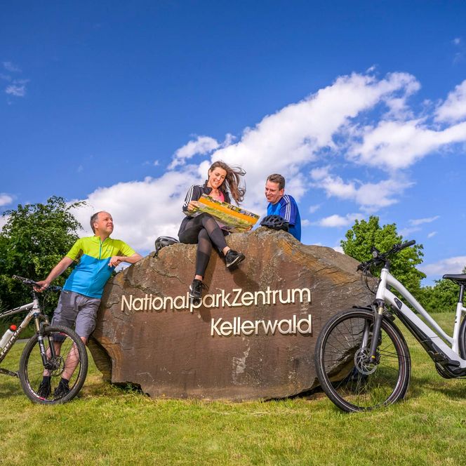 Drei Radler gruppieren sich um und auf den Begrüßungsstein am Nationalparkzentrum Vöhl-Herzhausen
