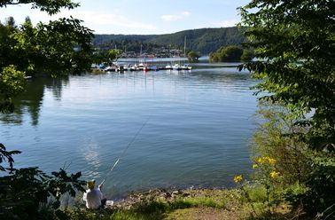 Ein Angler angelt im Sommer am Ufer des Edersees. Im Hintergrund ein Bootsanleger .