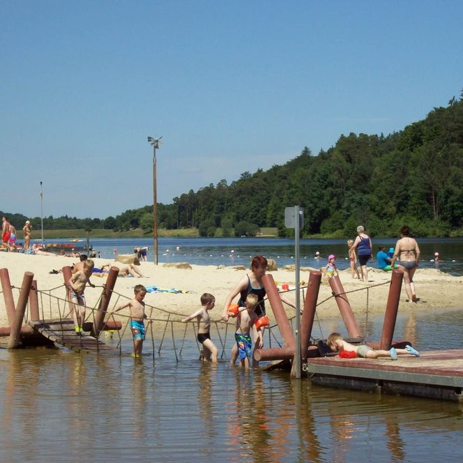 Der Strand des Strandbades am Twistesee im Sommer mit einigen Badegästen, die über eine kleine Wasserbrücke gehen.