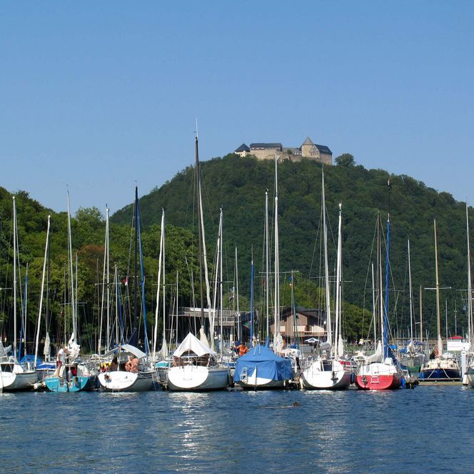 Im Vordergrund Bootsanleger mit Segelbooten, im Hintergrund thront Schloss Waldeck auf dem begrünten Hausberg