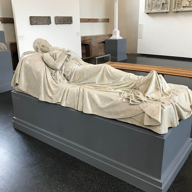 Eine cremefarbene, liegende Statuette der Königin Luise von Preußen von C. D. Rauch. Die Hände sind gefaltet. Im Hintergrund verschiedene Skulpturen, die im Museum ausgestellt werden.