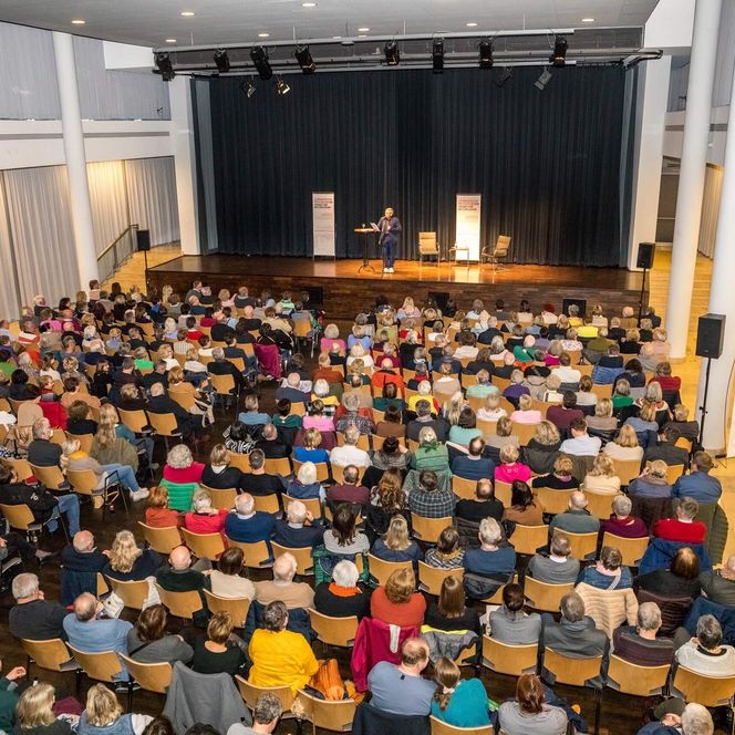 Ein Saal mit vielen sitzenden Zuschauern. Auf der Bühne steht ein Mann.