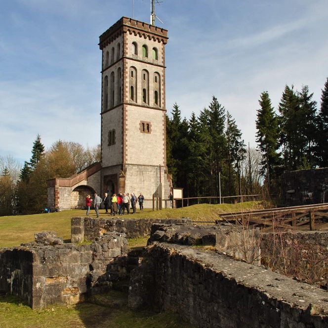 Das Bild zeigt einen hohen, rechteckigen Turm aus hellem Stein, der auf einem Hügel steht. Um den Turm herum befinden sich Bäume und Wiesen, und im Vordergrund sind Ruinenmauern aus Stein zu sehen. 