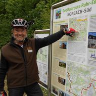 Ein Geoparkführer mit Fahrradhelm und Fahrradkleidung steht vor einer Infotafel und zeigt auf eine Karte.