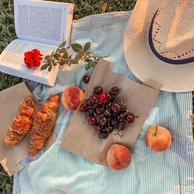 Auf einer hellen Decke liegen zwei Croissants, Kirschen auf einer braunen Serviette, ein aufgeklapptes Buch und ein Hut.