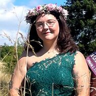 Eine junge Frau mit einem grünen Kleid und einem Blütenkranz steht in der grünen Natur und lächelt in die Kamera