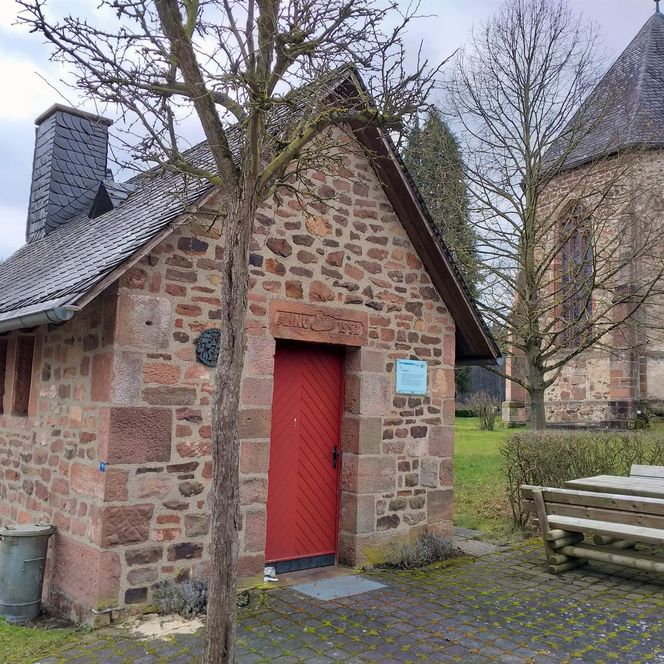 Im Vordergrund das aus Feldsteinen gemauerte Backhaus mit roter Tür. Im Hintergrund eine gotische Kirche und ein kahler Baum.