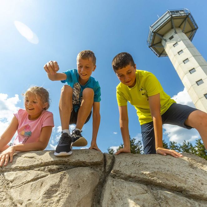 Mehrere Kinder stehen bei einem sonnigen Himmel fröhlich auf einem Stein vor dem Hochheideturm in Willingen