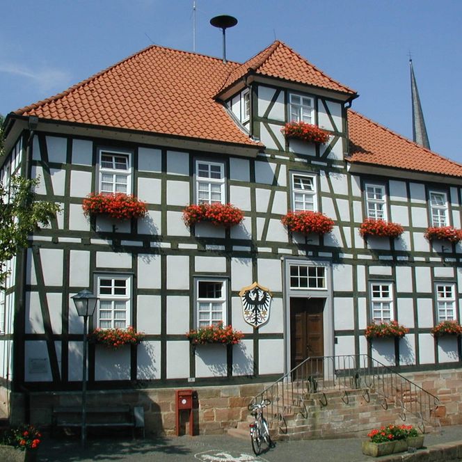 Ein zweistöckiges Fachwerkhaus mit beidseitigem Treppenaufgang. Vor den Fenstern hängen rote Geranien. Im Hintergrund der Kirchturm der Stadt.