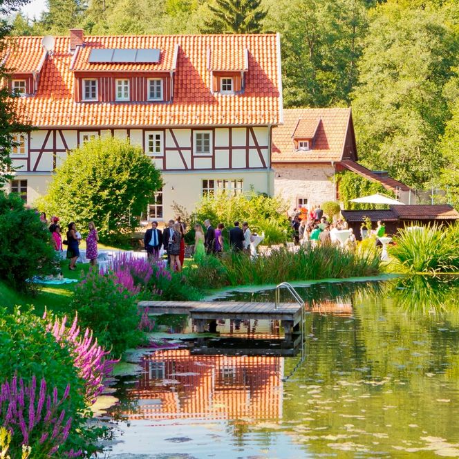 Aussenaufnahme des Landhauses bei einer Feierlichkeit. Im Vordergrund ein Teich, in der Aussenanlage halten sich einige festlich gekleidete Menschen auf.