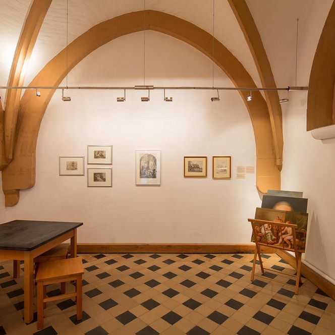 Ein Raum mit Tisch und Hockern links im Bild, an der Wand und rechts im Bild sind einige Bilder ausgestellt.