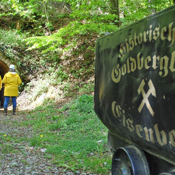 Einige Besucher mit gelben Jacken und Grubenhelm am Eingang des Bergwerks, im Vordergrund eine Lore