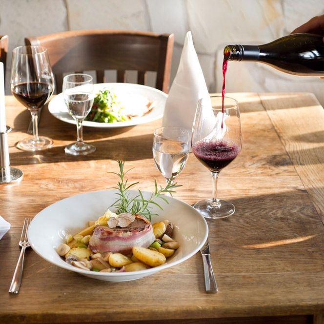 Ein gedeckter Tisch mit zwei Tellern, Besteck, Kerzen und Servietten. Ein Glas Rotwein wird eingeschenkt.