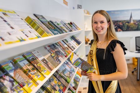 Emma Kuhlmann, die Korbacher Goldmarie, steht in der Tourist-Information vor einem Prospektregal. Sie trägt ein schwarzes Kleid mit goldener Schärpe und hält einen Flyer in der Hand.