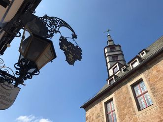 Links im Bild sieht man ein historisches Ladenschild eines Hotels und rechts im Bild das Dach mit einem Turm vom historischen Rathaus in Korbach. Der Himmel ist strahlend blau. 