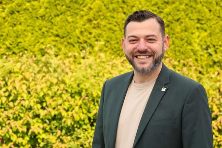 Dimitrij Schmalz steht vor einer grünen Hecke und lächelt in die Kamera.