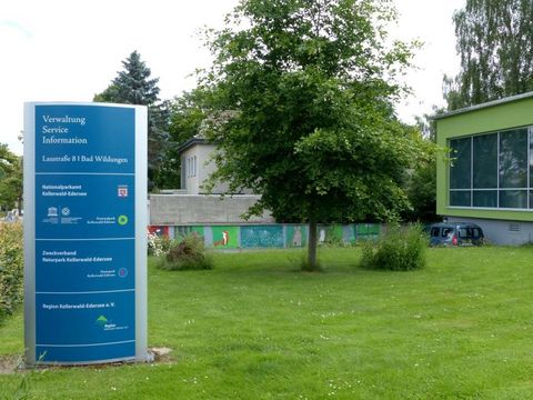 Eine blaue Infotafel mit den Verwaltungsstellen des Kellerwaldes steht auf einer grünen Wiese. Im Hintergrund eine kleine Mauer und ein Gebäudeteil