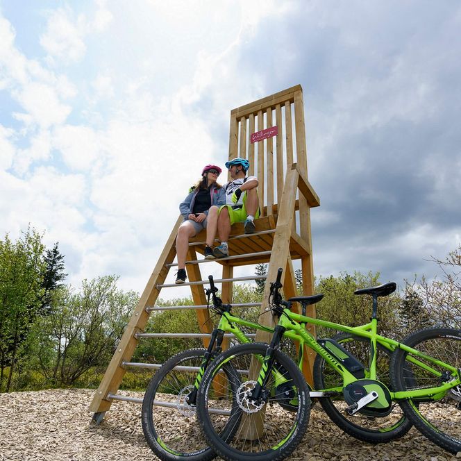 Ein Radfahrer und eine Radfahrerin sitzen auf dem hohen Stuhl, der am Ettelsberg steht. Die neongrünen Mountainbikes lehnen unten am Stuhl.